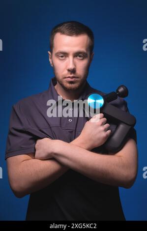 Ritratto di un massaggiatore professionista sicuro di sé con un massaggiatore a percussione in mano. Tasto basso, sfondo blu. Massaggio con onde d'urto Foto Stock