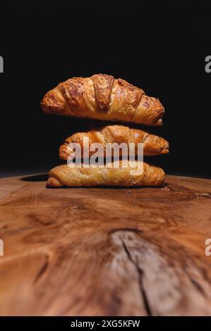 Primo piano di una pila di tre croissant su un asse di legno su sfondo scuro. Colazione deliziosa e salutare Foto Stock