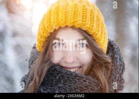 Ritratto ravvicinato di una giovane donna caucasica sorridente con un brillante cappello di lana e sciarpa, sullo sfondo di una foresta innevata Foto Stock