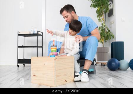 Giovane terapista che aiuta il ragazzo carino che ha paralisi cerebrale, giocando con lo sviluppo giocattolo in clinica di riabilitazione. Foto di alta qualità Foto Stock