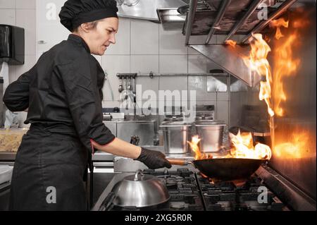 Chef donna che cucinava wok in cucina. Cuocere il wok fiammeggiante con verdure nella cucina commerciale. Fotografia di alta qualità Foto Stock