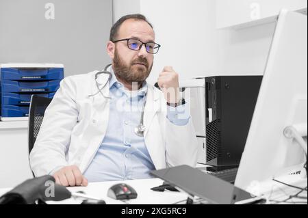 Ritratto di amichevole sorridente medico terapista seduto sul posto di lavoro. Buon medico sicuro maschio che indossa il cappotto medico bianco Foto Stock