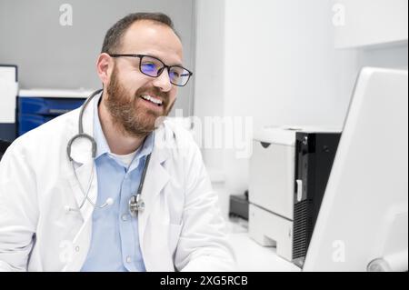 Ritratto di amichevole sorridente medico terapista seduto sul posto di lavoro. Buon medico sicuro maschio che indossa il cappotto medico bianco Foto Stock