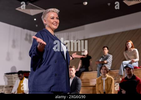 Docente femminile di abbigliamento formale sorridente, tenendo conferenze a gruppi di persone sullo sfondo, le persone si sono riunite per ascoltare parlare di impor Foto Stock