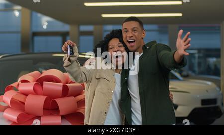 Eccitata giovane coppia felice in amore che celebra l'acquisto di una nuova auto regalo di lusso con un grande arco sul cofano dell'automobile donna afroamericana che mostra uomo Foto Stock