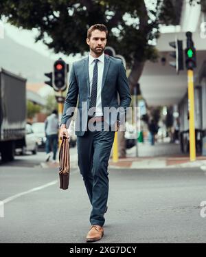 Uomo d'affari, valigetta e passeggiata lungo la strada in città accanto al semaforo, alla sicurezza e al cartello con le indicazioni per il lavoro. Persona, abito e borsa in pelle in metro Foto Stock