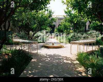 Sereno giardino con panche ornate e una tranquilla fontana d'acqua, circondato da vegetazione lussureggiante e alberi sotto la luce naturale del sole. Foto Stock