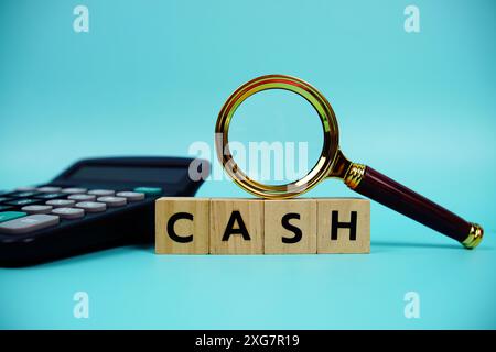 Contanti con blocchi di legno lettere alfabetiche, lente d'ingrandimento e calcolatrice su sfondo blu Foto Stock