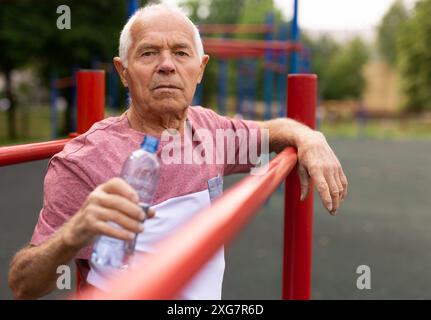 L'uomo anziano riposa e beve l'acqua da una bottiglia dopo aver fatto jogging nel parco Foto Stock