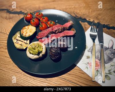 bistecca di fianco, manzo, verdure, media rara, grigliata, zucchine, pomodori ciliegini, funghi ripieni, cipolla caramellata, piatto nero, rustico, delizioso Foto Stock