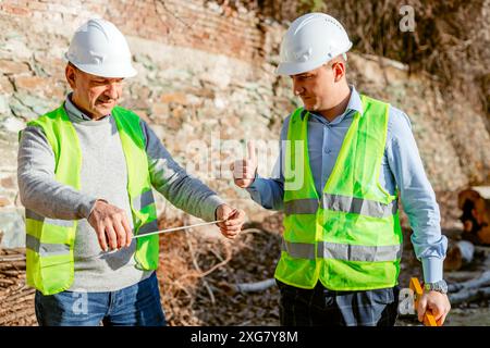 Due operai edili che indossano giubbotti di sicurezza e cappelli rigidi stanno misurando una distanza in un cantiere edile. Foto Stock