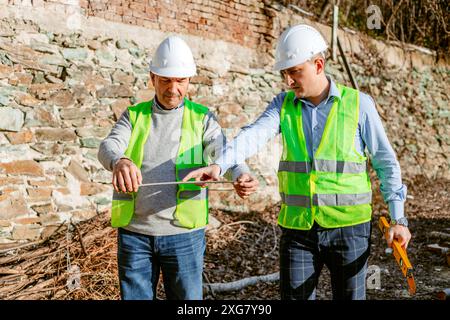 Due operai edili che indossano cappelli rigidi e giubbotti di sicurezza stanno lavorando in un cantiere. Il lavoratore più anziano ha un livello e il più giovane Wo Foto Stock