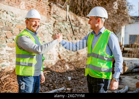 Due operai edili, che indossano cappelli rigidi bianchi e giubbotti di sicurezza gialli, stanno di fronte a una parete di mattoni e stringono la mano. Foto Stock