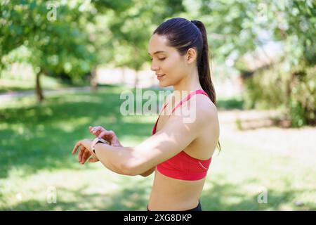 Una donna attiva nel parco utilizza uno smartwatch per tenere traccia del suo allenamento all'aperto, concentrandosi sul fitness e sul monitoraggio delle prestazioni Foto Stock