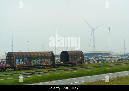 Gent, Belgio - 22 maggio 2023: Due vagoni merci recanti graffiti colorati sono parcheggiati sui binari, con una grande turbina eolica nel caos Foto Stock