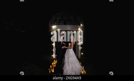 Gli sposi alla moda si stagliano di notte sullo sfondo di eleganti decorazioni e fiori, lampadine e ghirlande, in controluce. Un paio di spose Foto Stock