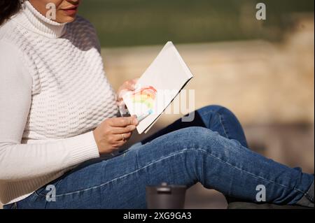 Primo piano di una donna che disegna un arcobaleno colorato in uno sketch book mentre siede all'aperto con un maglione bianco e un jeans casual. Foto Stock