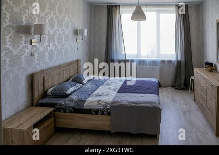 Interno di una piccola camera da letto. Camera da letto in un moderno appartamento con letto doppio con copriletto blu, comodino, cassettiera, due lampade da parete Foto Stock