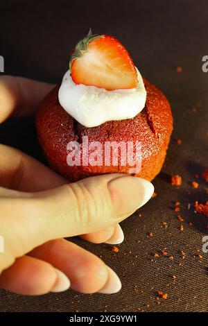 Tenere a mano un cupcake di velluto rosso ricoperto di panna e una fetta di fragola su sfondo scuro Foto Stock