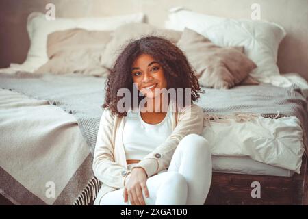 Allegro affascinante giovane afroamericana con capelli ricci in camera da letto, si siede sul pavimento ai piedi di fatto letto matrimoniale, guardando la macchina fotografica con ampio Foto Stock