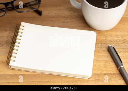 Modello a spirale per blocco note con tazza da caffè e occhiali su tavolo in legno. Carta bianca vuota Foto Stock