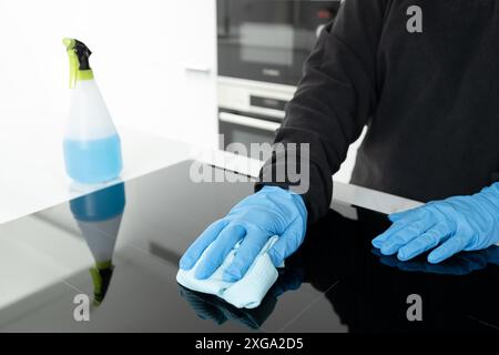 Mani in guanti protettivi pulizia di una stufa elettrica moderna con straccio di una cucina minimalista Foto Stock