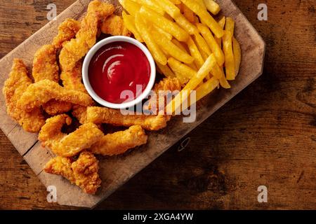 Aste di pollo, pezzi di carne disossati croccanti, con un tuffo rosso e patatine fritte, piatto sopra la testa sparare su uno sfondo di legno con un posto per il testo Foto Stock