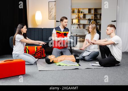 Istruttore di giovane uomo che aiuta a fare le compressioni di cuore di primo soccorso con il dummy durante l'addestramento di gruppo all'interno. Foto Stock