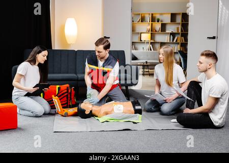 Istruttore di giovane uomo che aiuta a fare le compressioni di cuore di primo soccorso con il dummy durante l'addestramento di gruppo all'interno. Foto Stock