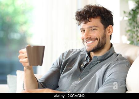 Uomo felice che tiene una tazza di caffè guardando la macchina fotografica in una casa Foto Stock