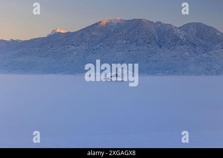 Capanna in legno al tramonto di fronte alle montagne, inverno, Loisach-Kochelsee-Moor, vista su Rabenkopf e Jochberg, Baviera, Germania, Europa Foto Stock