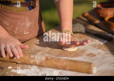Una donna sta facendo un impasto su un tavolo di legno. Sta indossando un grembiule marrone. L'impasto viene arrotolato con un mattarello Foto Stock