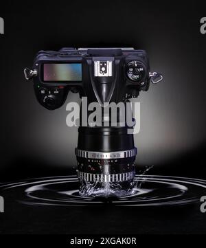 Fotocamera digitale nera con teleobiettivo immerso in acqua con increspature su sfondo scuro. Concetto di attrezzatura fotografica resistente alle intemperie. Foto Stock
