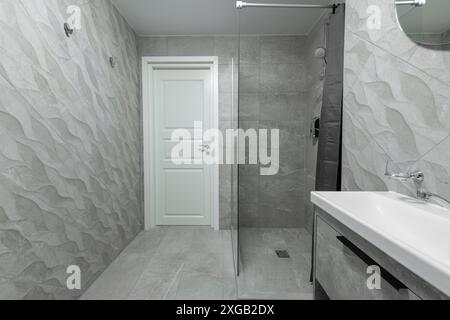 Bagno luminoso, elegante, moderno e minimalista. Design interno della doccia con porta bianca, pareti in mattonelle grigie, doccia, lavandino Foto Stock