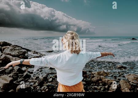 Una donna anziana che indossa una camicia bianca si allunga con le braccia aperte, di fronte al mare sotto un cielo nuvoloso e tempestoso, in piedi su una costa rocciosa. Foto Stock