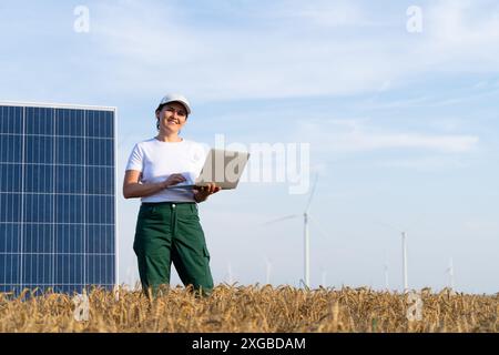 Donna agricoltrice che indossa un berretto bianco e una t-shirt con supporti per laptop accanto al pannello solare. Turbine eoliche sullo sfondo. Foto Stock
