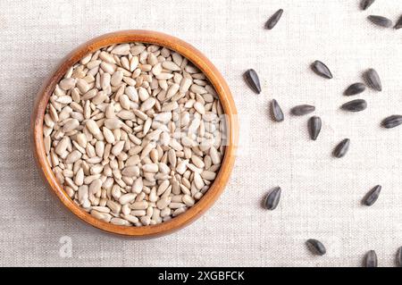 I semi di girasole sono stati sgusciati in una ciotola di legno su tessuto di lino. Frutti del seme oleoso Helianthus Annuus, il girasole comune. Sul lato destro semi interi. Foto Stock