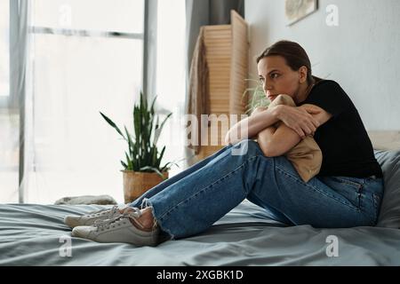 Una donna di mezza età in jeans e una t-shirt si siede tranquillamente sul letto di casa. Foto Stock