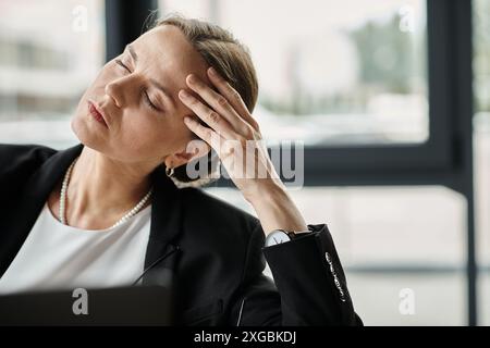 Una donna di mezza età in abito da lavoro tiene la testa mentre lavora con un notebook. Foto Stock