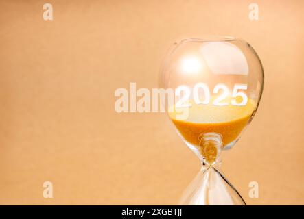 In arrivo per felice anno nuovo 2025, concetto di business. Primo piano il numero di calendario 3d 2025 anni affogato su sabbia dorata all'interno della clessidra isolata dalla luce ecologica Foto Stock