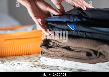 Una persona che piega i vestiti su un letto, con una valigia nelle vicinanze. Gli abiti includono camicie di colore scuro e asciugamani di colore marrone e bianco. Foto Stock