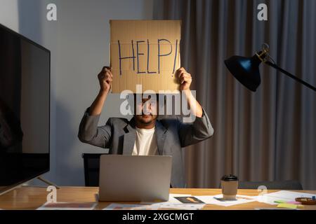 Uomo d'affari in tuta seduto alla scrivania dell'ufficio che lavora su un computer portatile chiedendo aiuto con un cartello di cartone. Un uomo d'affari disorientato regge una carta Foto Stock