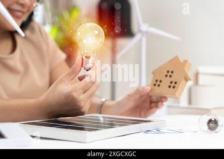 Donna con lampadina e mulino a vento. Vista ritagliata della donna che tiene la lampadina sul tavolo bianco con modello di turbina eolica, cella solare e casa a blocchi Foto Stock