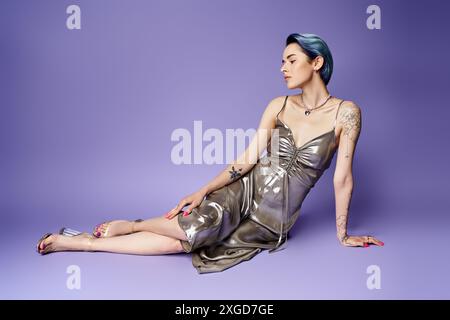 Giovane donna con i capelli corti blu, in abito da festa argento, seduta graziosamente sul pavimento in un ambiente studio. Foto Stock