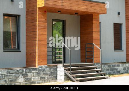 Una casa moderna esterna caratterizzata da un design elegante con rivestimenti in legno, accenti in pietra, una porta in vetro con telaio nero, scalini in cemento con ringhiera in metallo Foto Stock