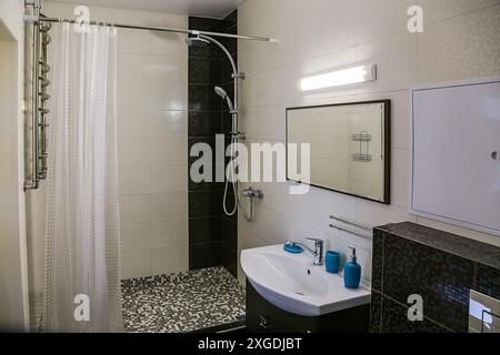 Interno di un bagno moderno e minimalista. Il lavandino si trova sotto lo specchio, la lampada sopra lo specchio, i saponi blu sul lavandino e il box doccia con cur Foto Stock