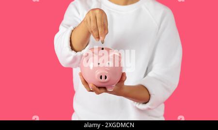 Persona che mette la moneta in Pink Piggy Bank su sfondo rosa Foto Stock