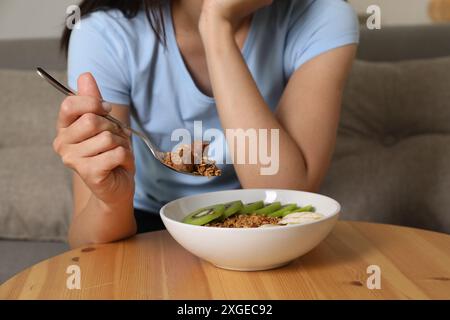 Disturbo alimentare. Donna che tiene un cucchiaio con granola sopra il recipiente al tavolo all'interno, primo piano Foto Stock