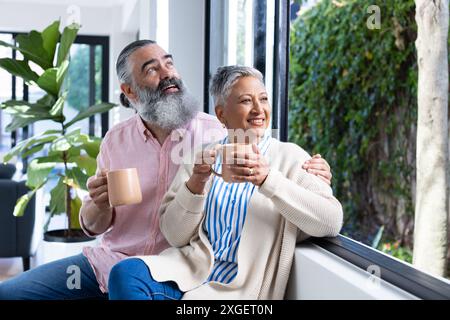 Una coppia di anziani sorridente si gusta un caffè insieme mentre guarda fuori dalla finestra di casa Foto Stock