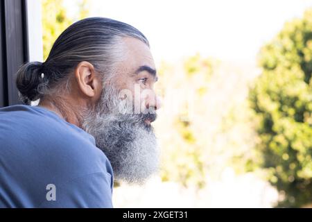 Uomo anziano con barba che guarda fuori dalla finestra, godendo momenti tranquilli, copiando spazio Foto Stock